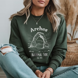 May include: Dark green sweatshirt featuring a white graphic of a rock arch with the text "Arches National Park - Est. 1971." The sweatshirt is a crew neck style and is made of a soft material. The overall theme is travel and national parks.