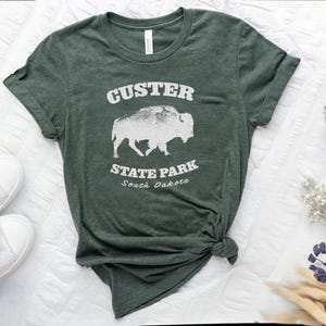 Image shows  a Heather forest green t-shirt with the text "Custer State Park" in white and a bison graphic. The shirt looks great  paired with jeans for a casual look. This athletic style tee is made of soft and cozy cotton and features the Bella and Canvas flattering fit and durability. These shirts are short sleeve crew neck style and make great adventure souvenirs for a hiking or camping trip to South Dakota as well as birthday, anniversary, Christmas, Mothers and Fathers Day gifts.