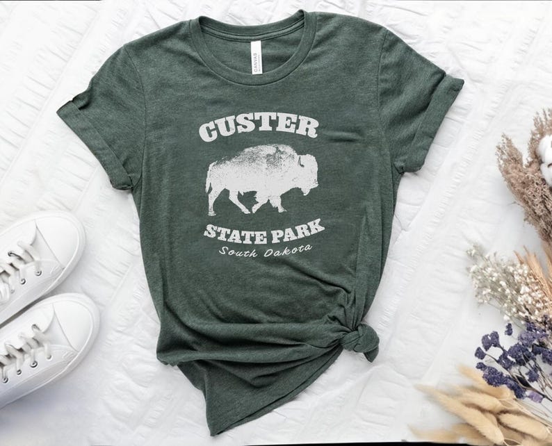 Image shows  a Heather forest green t-shirt with the text "Custer State Park" in white and a bison graphic. The shirt looks great  paired with jeans for a casual look. This athletic style tee is made of soft and cozy cotton and features the Bella and Canvas flattering fit and durability. These shirts are short sleeve crew neck style and make great adventure souvenirs for a hiking or camping trip to South Dakota as well as birthday, anniversary, Christmas, Mothers and Fathers Day gifts.