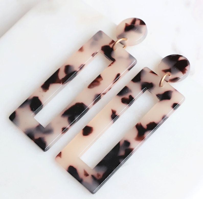 May include: A pair of statement earrings with a tortoiseshell pattern. The earrings are rectangular with a smaller rectangle cut out in the center. The earrings are made of a lightweight plastic material.