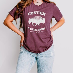 Pu&ograve; includere: T-shirt bordeaux con la scritta "Custer State Park South Dakota" e una grafica di un bisonte bianco. La maglietta ha maniche corte e girocollo. La modella indossa jeans azzurri.