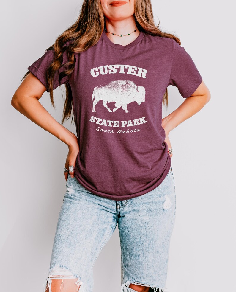 Pu&ograve; includere: T-shirt bordeaux con la scritta "Custer State Park South Dakota" e una grafica di un bisonte bianco. La maglietta ha maniche corte e girocollo. La modella indossa jeans azzurri.