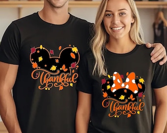May include: Two black t-shirts with a Thanksgiving themed design. The design features a Mickey Mouse head with fall leaves and the word "Thankful". One shirt has a bow on the Mickey Mouse head, while the other does not.