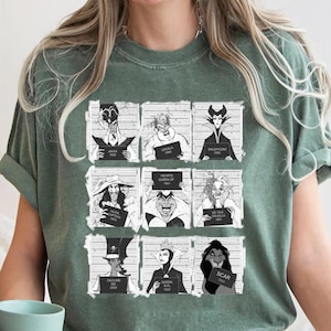 Puede incluir: Camiseta verde con una cuadr&iacute;cula de nueve villanos de dibujos animados al estilo de una foto policial. Cada villano sostiene un letrero con su nombre y un a&ntilde;o. La camiseta tiene un corte holgado y mangas cortas.