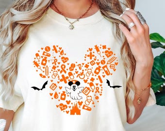 May include: Cream-colored t-shirt featuring a Mickey Mouse-shaped design filled with orange medical-themed icons, a ghost, and bats. The design includes pills, hearts, and syringes. The shirt is short-sleeved and has a crew neck.
