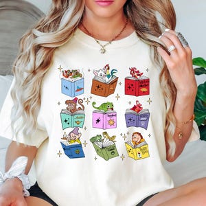 May include: Cream-colored t-shirt featuring colorful illustrations of open books with various cartoon characters. The books are in different colors and have gold star accents. The shirt is a casual, comfortable style.
