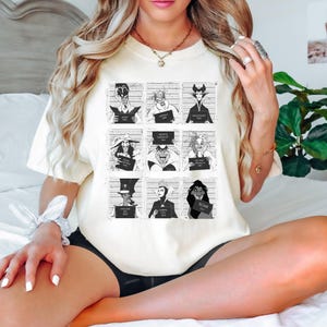 Puede incluir: Camiseta color crema con una cuadr&iacute;cula de ilustraciones en blanco y negro de villanos de dibujos animados en poses de foto policial. Cada personaje sostiene un letrero con un nombre y un n&uacute;mero. La camiseta tiene cuello redondo y mangas cortas.