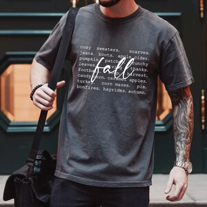 Może przedstawiać: Szary T-shirt z białym nadrukiem graficznym, na kt&oacute;rym widnieje napis "przytulne swetry, szaliki, dżinsy, buty, cydr jabłkowy, pole dyni, liście, jesień, chrupiący, dzięki, piłka nożna, zbiory, cukrowe kukurydza, jabłka karmelowe, indyk, labirynty kukurydziane, ciasto, ogniska, przejażdżki bryczką, jesień."