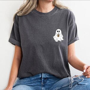Pu&ograve; includere: Una maglietta grigia scura con un fantasma bianco che tiene una tazza di caff&egrave;. Il fantasma ha due occhi e un piccolo sorriso.