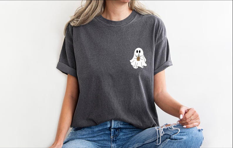 Pu&ograve; includere: Una maglietta grigia scura con un fantasma bianco che tiene una tazza di caff&egrave;. Il fantasma ha due occhi e un piccolo sorriso.