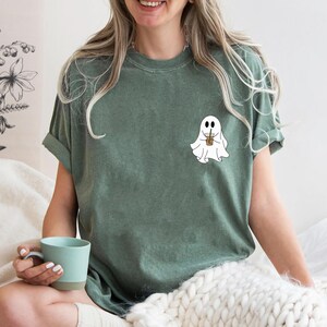 Pu&ograve; includere: Una maglietta verde con un fantasma bianco che tiene una bevanda marrone. Il fantasma ha un viso amichevole e indossa una cannuccia.