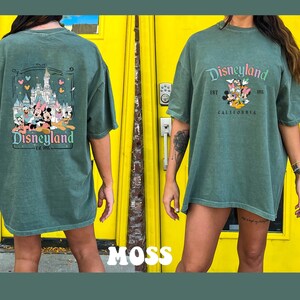 May include: A green oversized t-shirt with a vintage Disneyland graphic featuring Mickey Mouse and friends in front of Sleeping Beauty's Castle. The text reads "Disneyland Est. 1955 California".