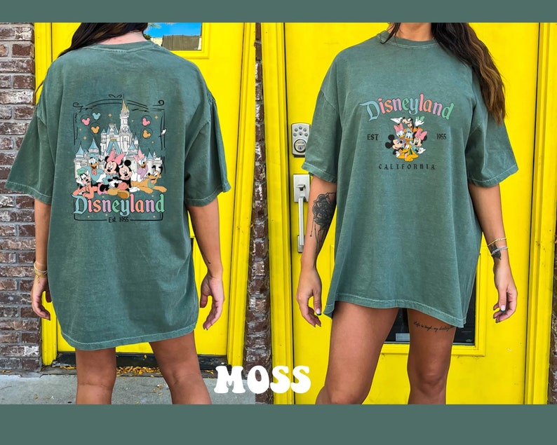 May include: A green oversized t-shirt with a vintage Disneyland graphic featuring Mickey Mouse and friends in front of Sleeping Beauty's Castle. The text reads "Disneyland Est. 1955 California".