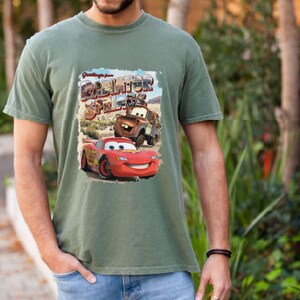 May include: Green t-shirt with a vintage-style graphic of Lightning McQueen and Mater from the Disney Pixar movie Cars. The graphic says "Greetings from Radiator Springs".