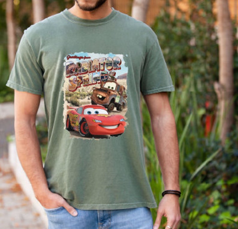 May include: Green t-shirt with a vintage-style graphic of Lightning McQueen and Mater from the Disney Pixar movie Cars. The graphic says "Greetings from Radiator Springs".