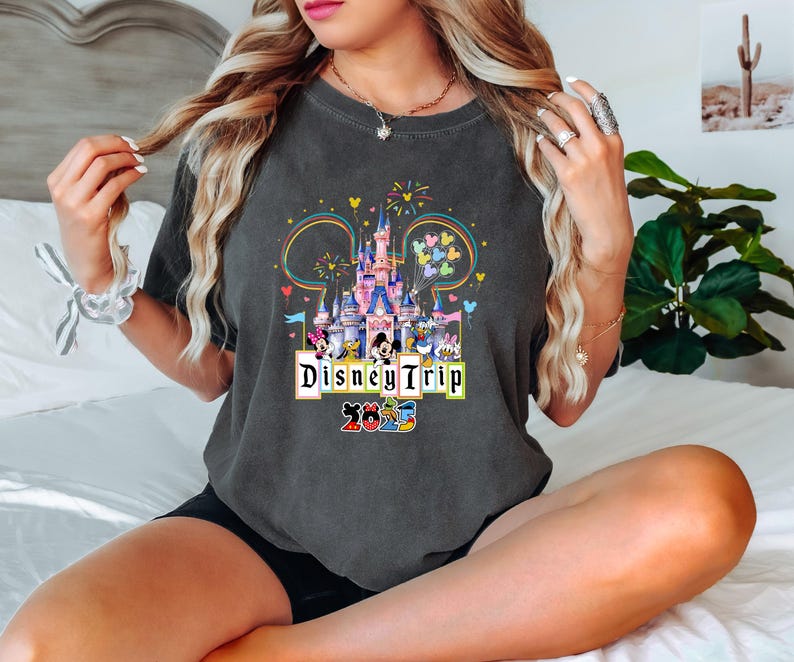 May include: A dark gray t-shirt with a colorful graphic of Cinderella's castle and Mickey Mouse characters. The text "Disney Trip 2023" is printed on the shirt.