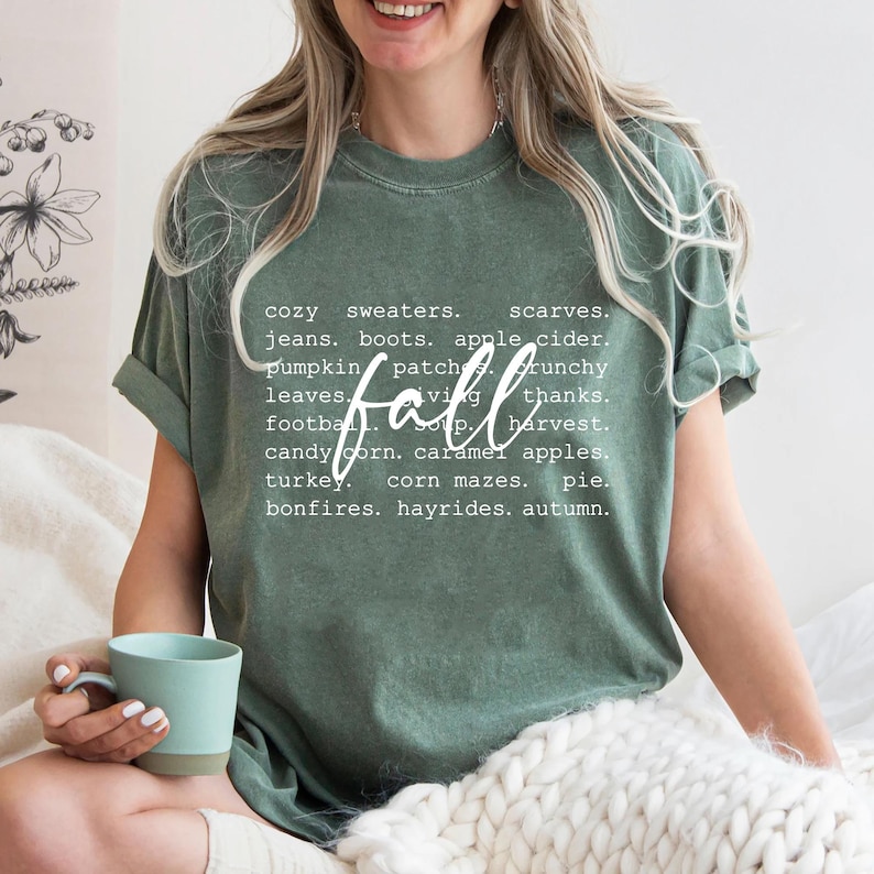 Może przedstawiać: Zielony T-shirt z białym nadrukiem, na kt&oacute;rym widnieje napis "fall" oraz lista sł&oacute;w związanych z porą roku, takich jak "przytulne swetry", "szaliki", "jabłkowy cydr", "pola z dyniami", "liście", "ogniska" i "przejażdżki na wozie z sianem".