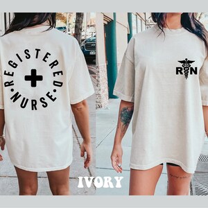 May include: A white oversized t-shirt with a black graphic on the back that says "Registered Nurse" in a circle with a plus sign in the center. The front of the shirt has a black graphic of a caduceus with the letters "RN" below it.