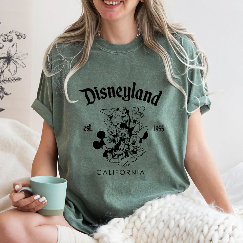 May include: Green t-shirt with a black graphic of Mickey Mouse and friends. The graphic says "Disneyland est. 1955 California".