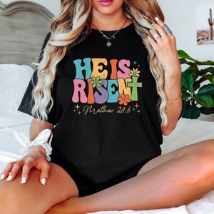 May include: A black t-shirt with a colorful retro-style graphic that says "He is Risen" with flowers and stars. The text "Matthew 28:6" is below the graphic.
