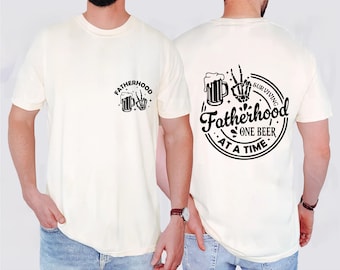 Può includere: Due t-shirt bianche con una stampa grafica nera di una birra e una mano scheletrica con il testo "Fatherhood Surviving One Beer At A Time".