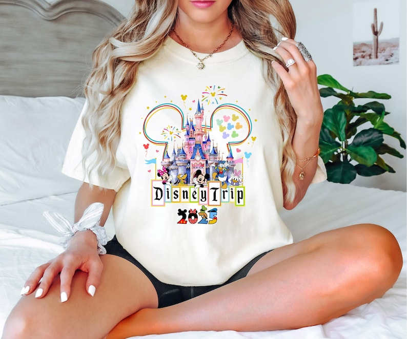 May include: A white t-shirt with a colorful graphic of Cinderella's castle and Mickey Mouse characters. The text "Disney Trip 2023" is printed below the graphic.