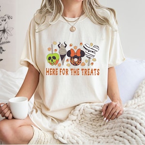 May include: A white t-shirt with a graphic of Halloween treats, including a green apple, an ice cream cone, a donut, and a Mickey Mouse-shaped lollipop. The text "Here for the treats" is printed below the graphic.