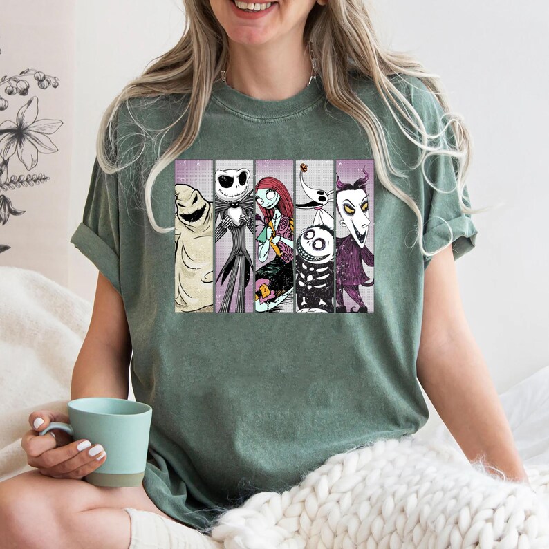 May include: Sage green t-shirt featuring a graphic of characters from The Nightmare Before Christmas. The design includes images of Oogie Boogie, Jack Skellington, Sally, Zero, and others. The shirt is a short-sleeved crew neck.