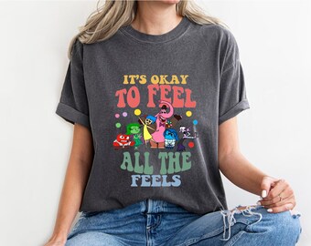 Könnte beinhalten: Ein dunkelgraues T-Shirt mit einer bunten Grafik von Figuren aus dem Film Inside Out. Der Text auf dem Shirt lautet "Es ist okay, alle Gefühle zu fühlen."