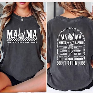 May include: Dark gray t-shirt with the text "MAMA" in white, featuring a skeleton hand gesture. The shirt also includes "THE MOTHERHOOD TOUR" and a list of activities. The back of the shirt has the text "MA MA MAKES IT ALL HAPPEN".