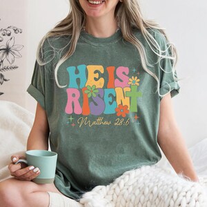 May include: A green t-shirt with a retro-style graphic that says "He is Risen" with flowers and stars. The text "Matthew 28:6" is below the graphic.