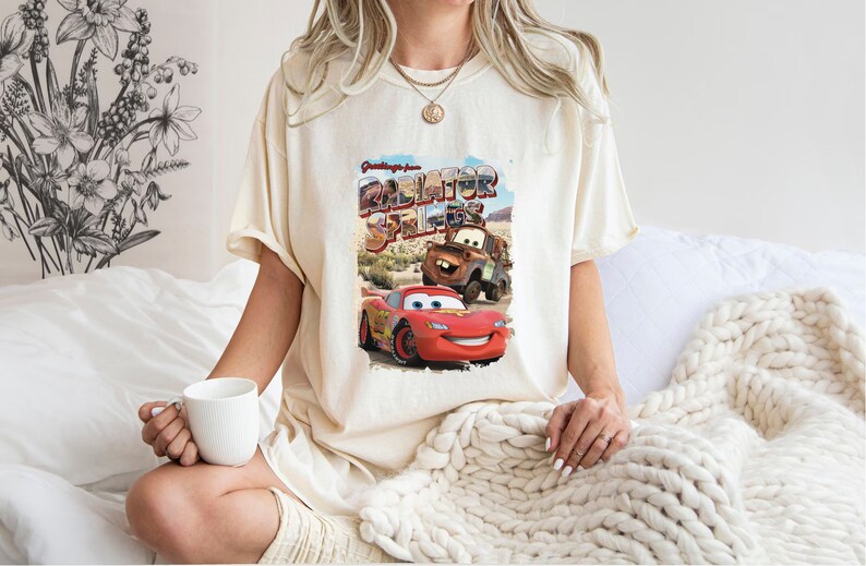 May include: A white t-shirt with a graphic print of Lightning McQueen and Mater from the movie Cars. The graphic says "Greetings from Radiator Springs".