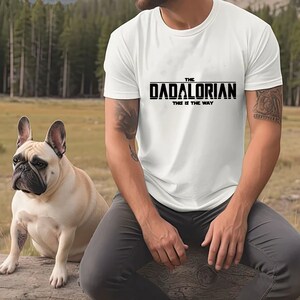 May include: White t-shirt with black text that reads "The Dadalorian This is the Way". A person wearing the shirt is sitting on a log with a French bulldog sitting next to them.