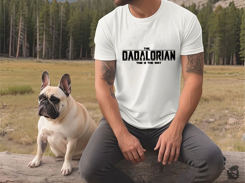 May include: White t-shirt with black text that reads "The Dadalorian This is the Way". A person wearing the shirt is sitting on a log with a French bulldog sitting next to them.