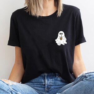 Pu&ograve; includere: Una maglietta nera con un fantasma bianco che tiene una tazza di caff&egrave;. Il fantasma ha due occhi e un piccolo sorriso.