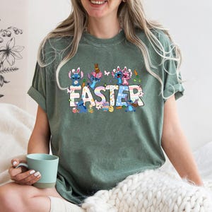 May include: Green short-sleeved t-shirt with a colorful graphic of Stitch characters celebrating Easter. The graphic includes the word "Easter" in colorful letters.