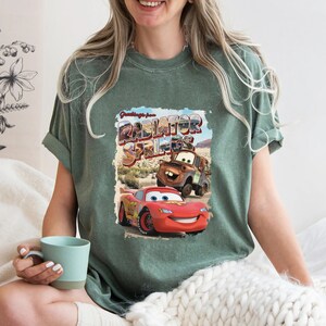 May include: A green t-shirt with a vintage postcard design featuring the text "Greetings from Radiator Springs" and images of Lightning McQueen and Mater from the Disney Pixar Cars movie.