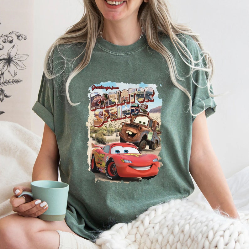 May include: A green t-shirt with a vintage postcard design featuring the text "Greetings from Radiator Springs" and images of Lightning McQueen and Mater from the Disney Pixar Cars movie.