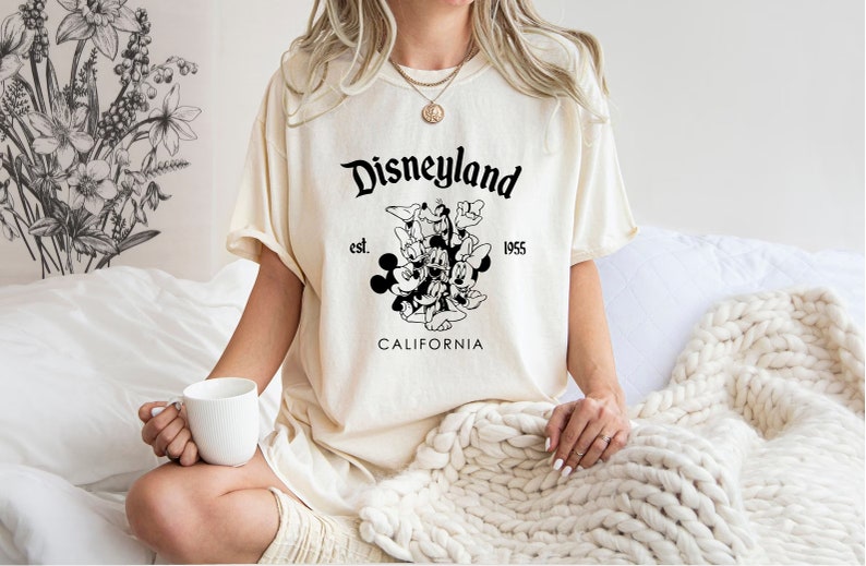 May include: A white t-shirt with a black graphic of Mickey Mouse and friends. The graphic includes the text "Disneyland est. 1955 California".