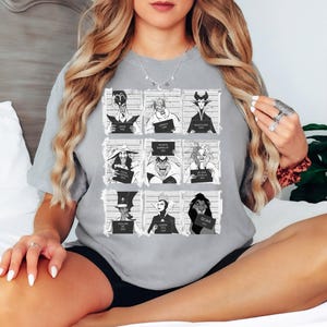 Puede incluir: Una camiseta gris con una cuadr&iacute;cula de nueve ilustraciones en blanco y negro de villanos de Disney. Cada villano sostiene un letrero con su nombre y un a&ntilde;o. La camiseta es de estilo casual y c&oacute;modo.