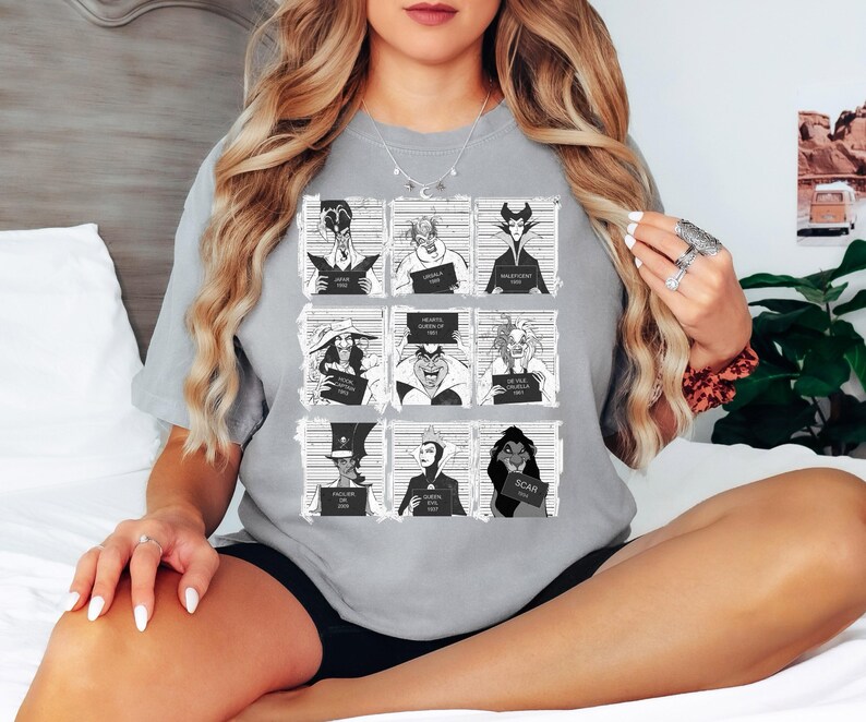 Puede incluir: Una camiseta gris con una cuadr&iacute;cula de nueve ilustraciones en blanco y negro de villanos de Disney. Cada villano sostiene un letrero con su nombre y un a&ntilde;o. La camiseta es de estilo casual y c&oacute;modo.