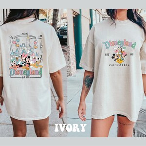 May include: A white oversized t-shirt with a graphic print of Mickey Mouse and friends in front of the Disneyland castle. The text "Disneyland Est. 1955 California" is printed on the front of the shirt. The back of the shirt has a similar graphic print with the text "Disneyland Est. 1955".