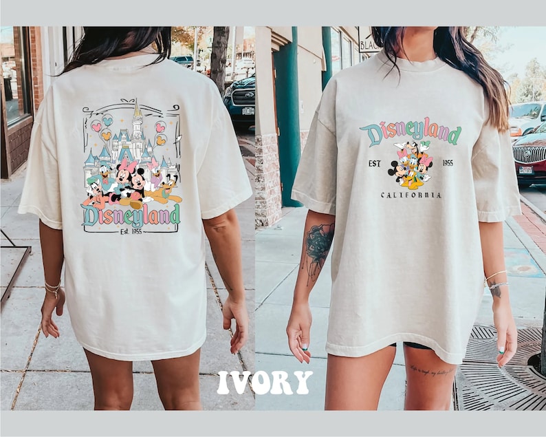 May include: A white oversized t-shirt with a graphic print of Mickey Mouse and friends in front of the Disneyland castle. The text "Disneyland Est. 1955 California" is printed on the front of the shirt. The back of the shirt has a similar graphic print with the text "Disneyland Est. 1955".