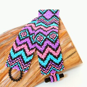May include: Two beaded bracelets with a geometric pattern in pink, blue, purple, and black. The bracelets are lying on a piece of wood.