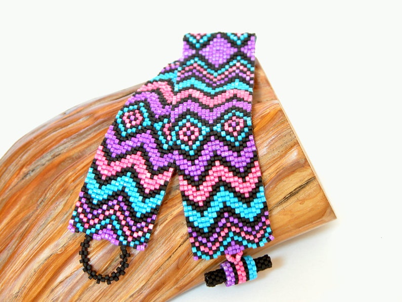 May include: Two beaded bracelets with a geometric pattern in pink, blue, purple, and black. The bracelets are lying on a piece of wood.