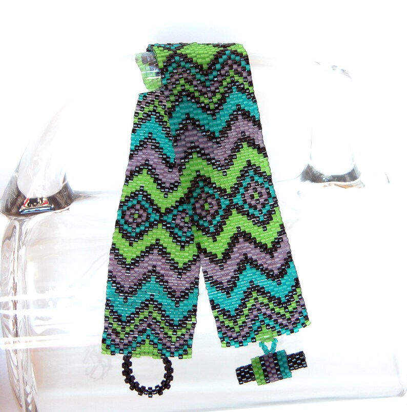 May include: Two beaded bracelets with a chevron pattern in shades of green, blue, black, and purple. The bracelets are tied together with a black cord and a small beaded charm.