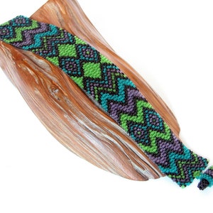 May include: A green, purple, and black beaded bracelet with a geometric pattern. The bracelet is on a wooden surface.
