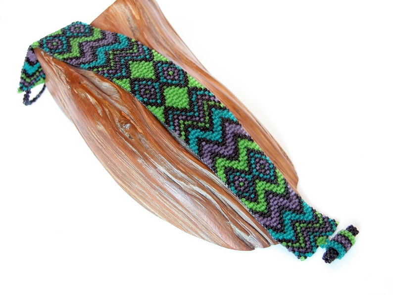 May include: A green, purple, and black beaded bracelet with a geometric pattern. The bracelet is on a wooden surface.