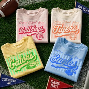 Peut inclure: Quatre t-shirts en rose clair, crème, jaune et bleu clair, chacun présentant des rayures verticales, le texte Custom School Name, et les noms d'équipe Bulldogs, Tigers, Gators ou Eagles avec des graphismes d'animaux assortis.