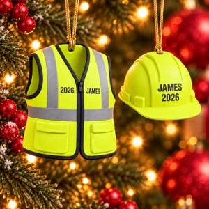 May include: A neon yellow high-visibility safety vest ornament and a matching hard hat ornament, both hanging by gold cords. The vest features grey reflective stripes and the text "2026 JAMES". The hard hat displays the text "JAMES 2026".
